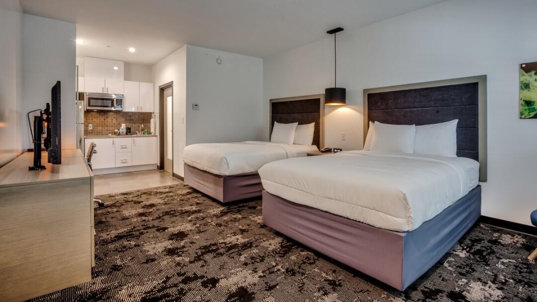 A wide view of a hotel room with two double beds, a kitchenette, and modern furnishings.