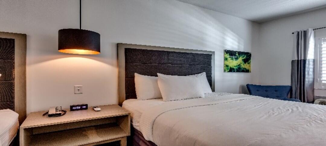 A hotel room with a bed, nightstand, lamp, chair, and artwork.