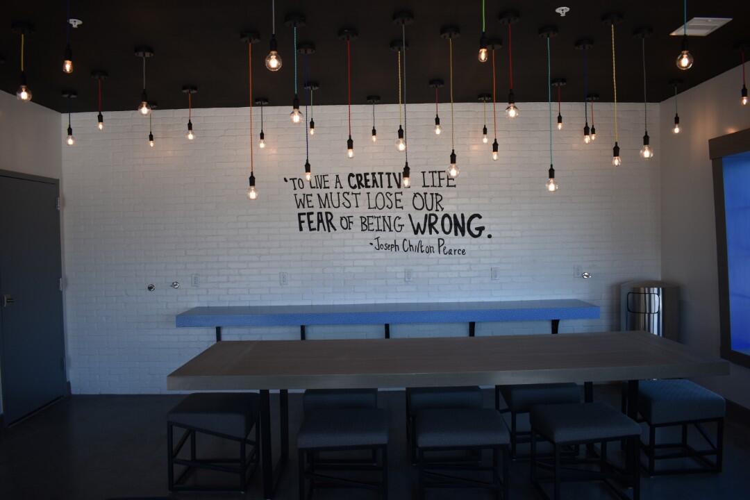A dining area with a large table and chairs, a quote on the wall, and hanging lights.